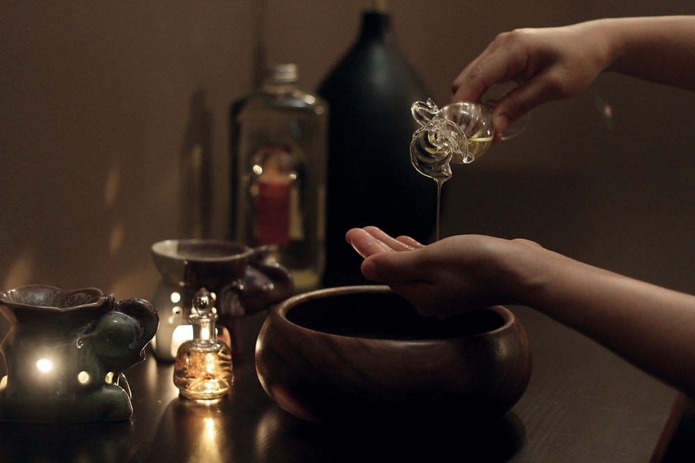 Hands pouring golden oil into a palm over a wooden bowl beside a lit elephant oil burner in a dim spa setting