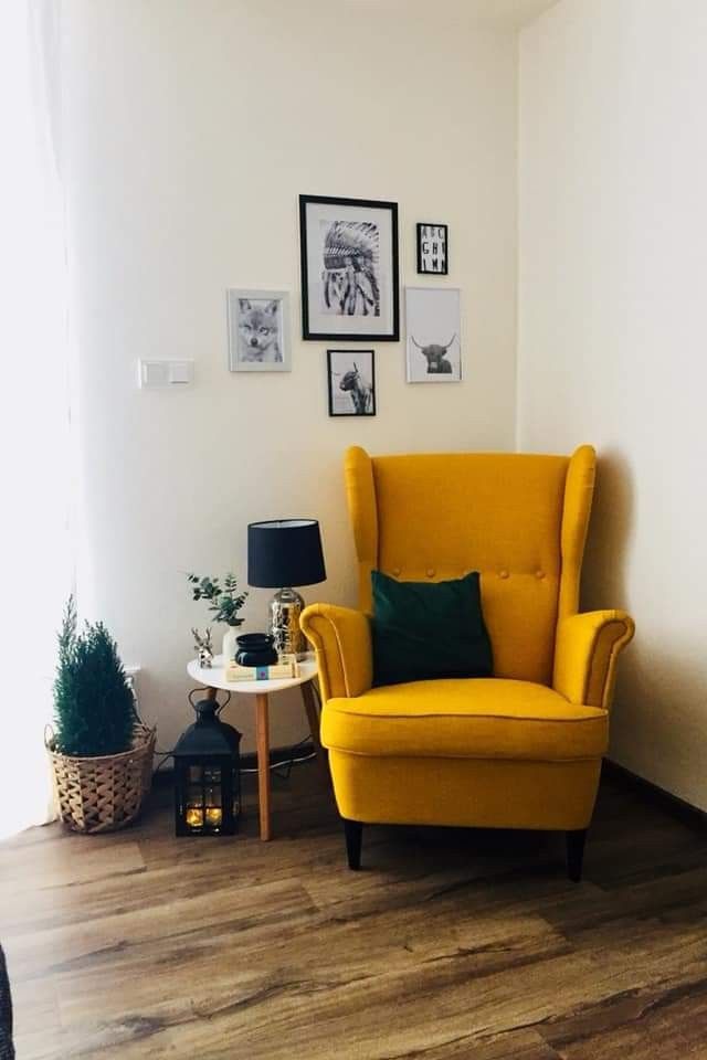 Cozy reading nook with a yellow wingback armchair, side table with lamp and books, potted plant, lantern, and framed wall art